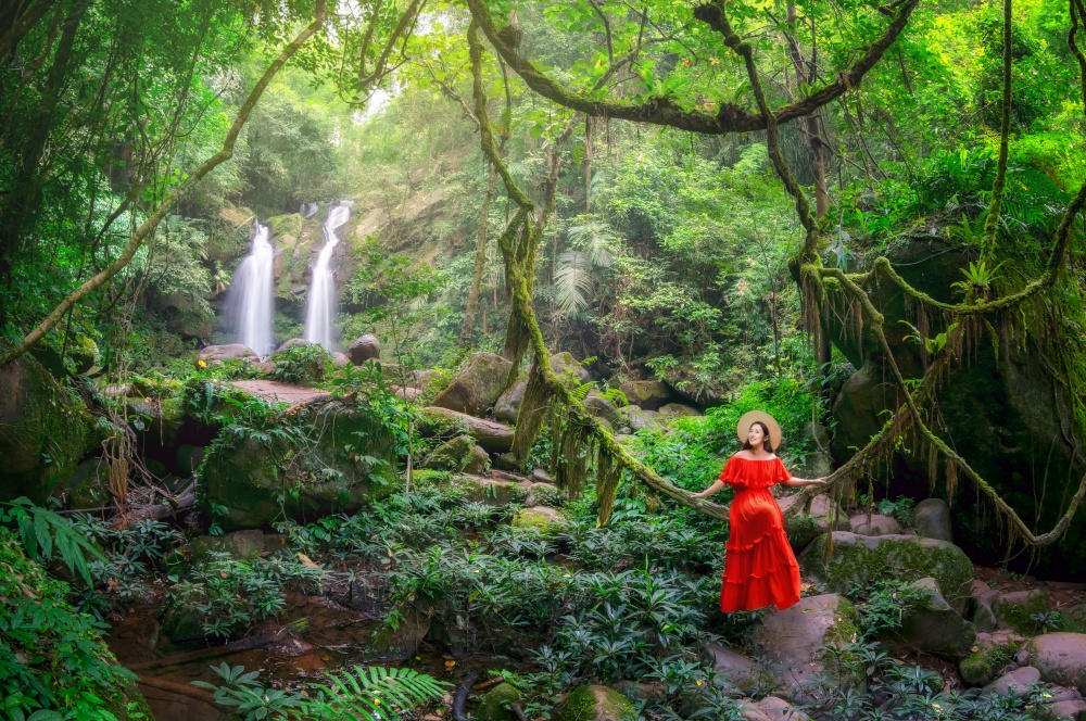 Beautiful woman in red dress sitting on the vine in front of the Sapan Waterfall, Khun Nan National Park, Boklua District, Nan Province, Thailand.. Sapan Waterfall