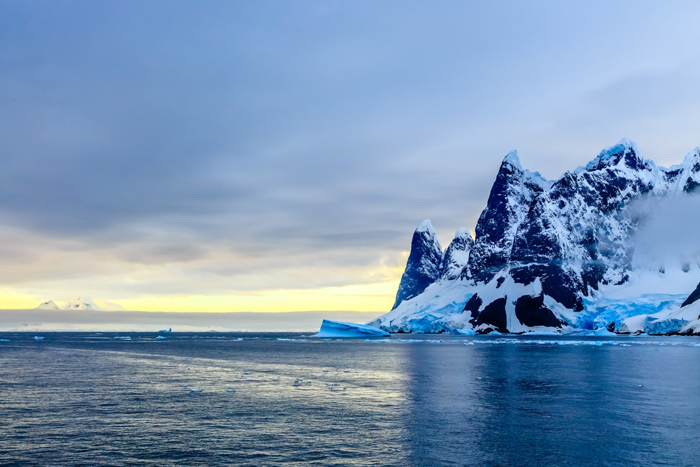 Sunset over cliffs, blue glacier and drifting iceberg with water surface in foreground, close to Argentine islands, Antarctica