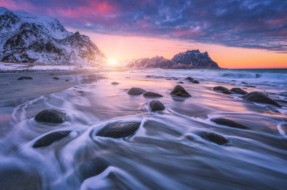 Sandy beach with stones in blurred water, colorful pink with blue clouds sky and snowy mountains at sunset. Utakleiv beach, Lofoten islands, Norway. Winter landscape with sea, waves, rocks at twilight