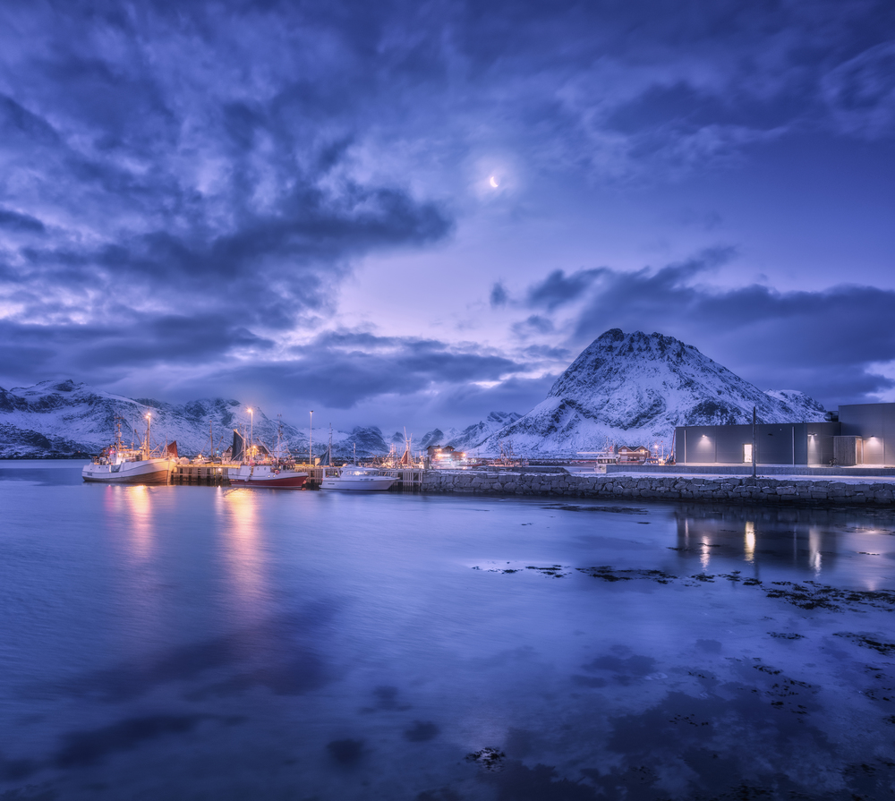 Fishing boats near pier on the sea against snowy mountains and starry sky with moon at night in Lofoten islands, Norway. Winter landscape with ship, buildings, illumination, high rocks and clouds