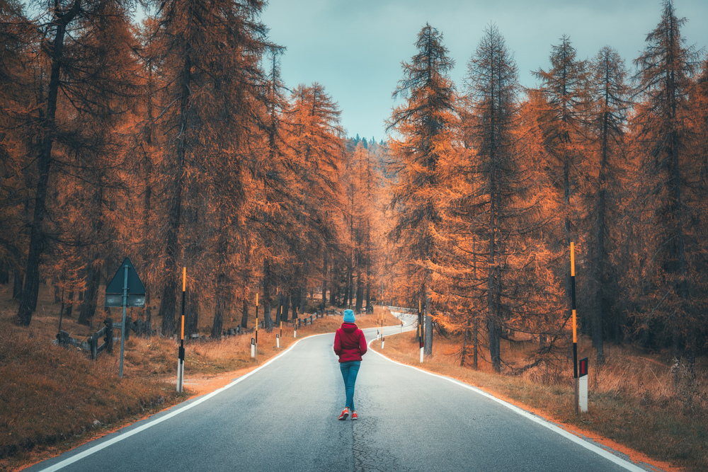 Young woman on the road in autumn forest at sunset. Landscape with girl in red jacket, mountain road, orange trees. Empty asphalt roadway through woodland in fall. Travel and adventure in Europe