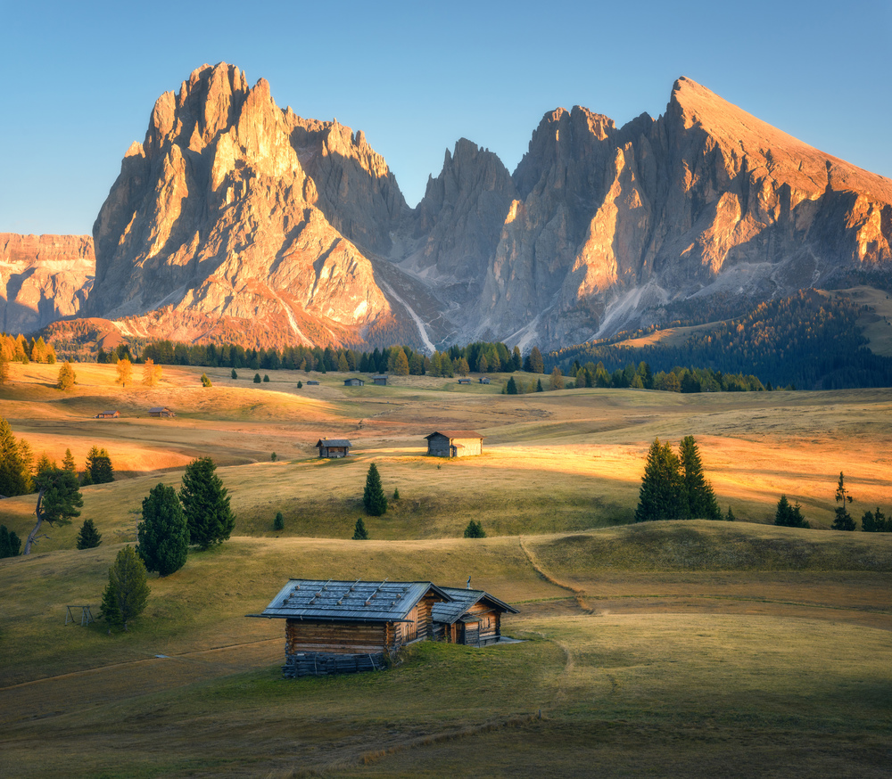 Small wooden houses in beautiful mountain valley at sunset. Autumn landscape with mountains, buildings, hill with green grass, trees, high rocks, sky. Alpe di Siusi in Dolomites, Italy. Italian alps