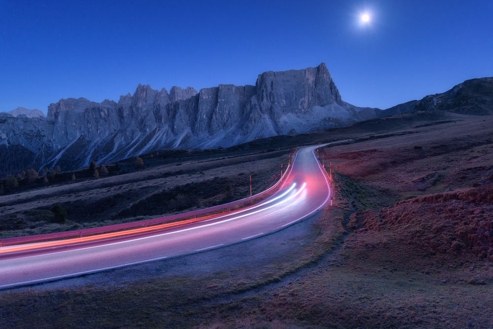 Blurred car headlights on winding road at night in autumn. Landscape with asphalt road, light trails, mountains, hills, blue sky with moonlight at dusk. Roadway in Italy. Moon over highway and rocks