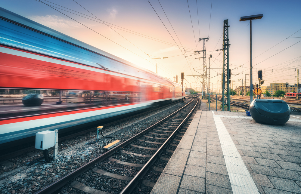 Red high speed train in motion on the railway station at colorful sunset. Blurred modern intercity train with sky reflection in windows on the railway platform. Passenger transportation. Railroad