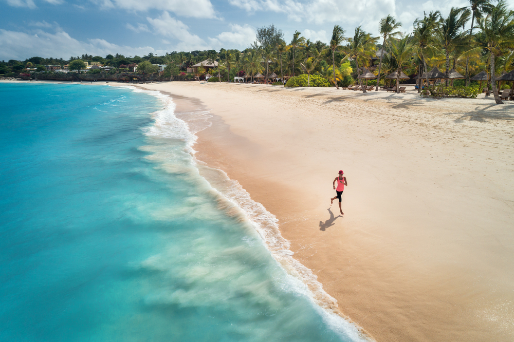 Aerial view of the running young woman on the white sandy beach near sea with waves at sunrise. Summer holiday. Top view of sporty slim girl, clear azure water. Indian Ocean. Lifestyle and sport