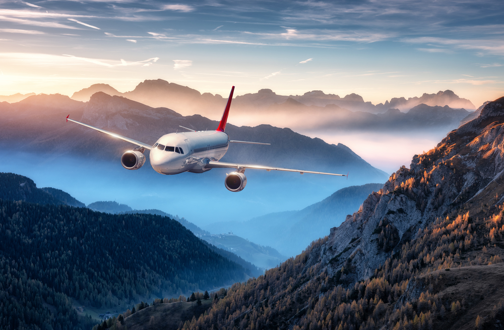 Airplane is flying over mountains in fog at colorful sunset in summer. Landscape with passenger airplane, hills in low clouds, blue sky. White aircraft. Business travel. Commercial plane. Aerial view