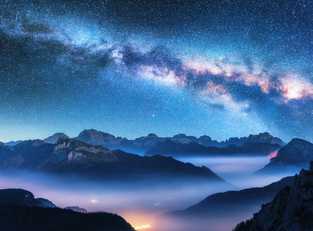 Milky Way above mountains in fog at night in summer. Landscape with alpine mountain valley, low clouds, purple starry sky with milky way, city illumination. Aerial. Passo Giau, Dolomites, Italy. Space