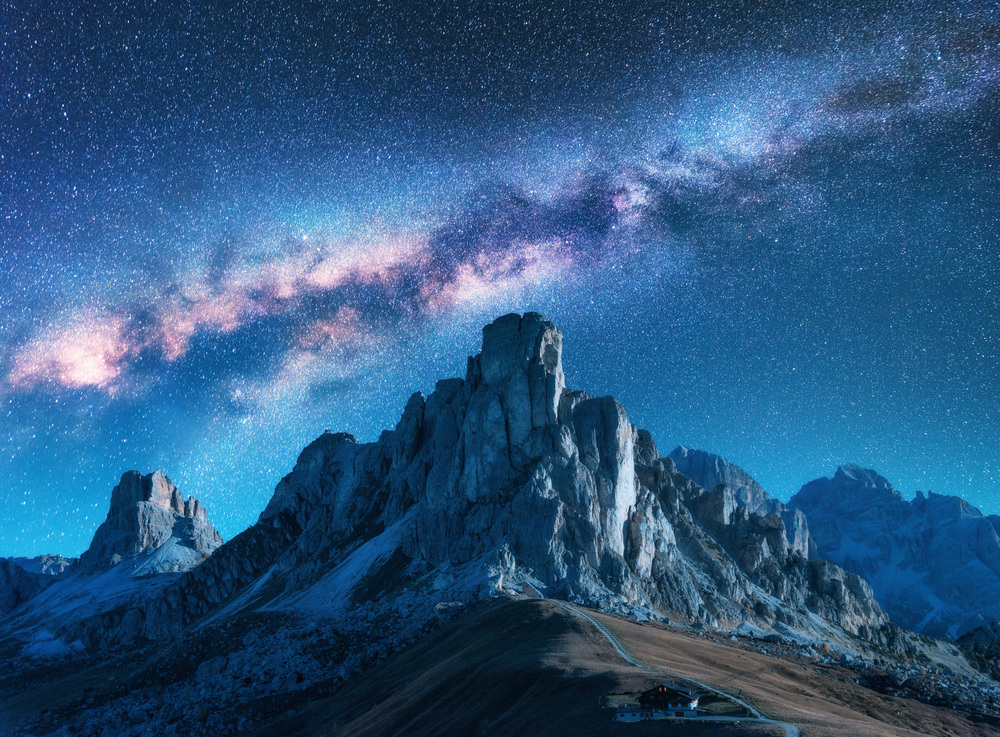 Milky Way above mountains at night in summer. Landscape with alpine mountain valley, blue sky with milky way and stars, buildings on the hill, rocks. Aerial view. Passo Giau in Dolomites, Italy. Space