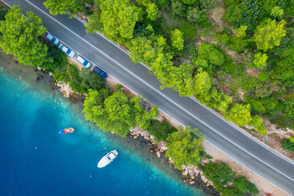 Aerial view of road in beautiful green forest and sea coast at sunset in spring. Colorful landscape with cars on roadway, boat, blue water, trees in summer. Top view of highway in Croatia. Travel