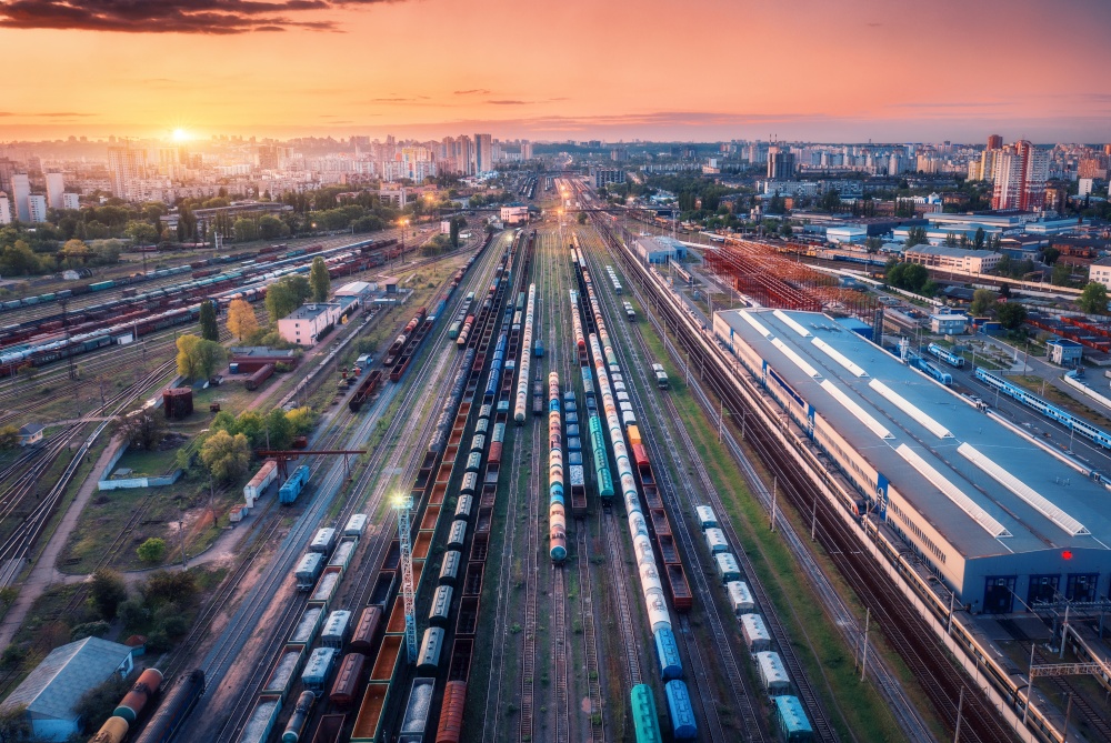 Aerial view of freight trains at sunset. Top view of railway station, wagons, railroad. Heavy industry. Landscape with train in depot, buildings, city, lights, colorful sky at night. Transportation