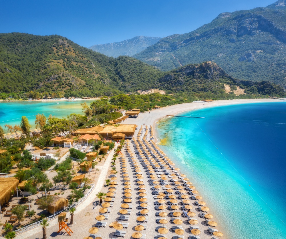 Aerial view of sea bay, sandy beach with umbrellas, palm trees, mountain at sunset in summer. Blue lagoon in Oludeniz, Turkey. Tropical landscape with island, white sandy bank, blue water. Top view