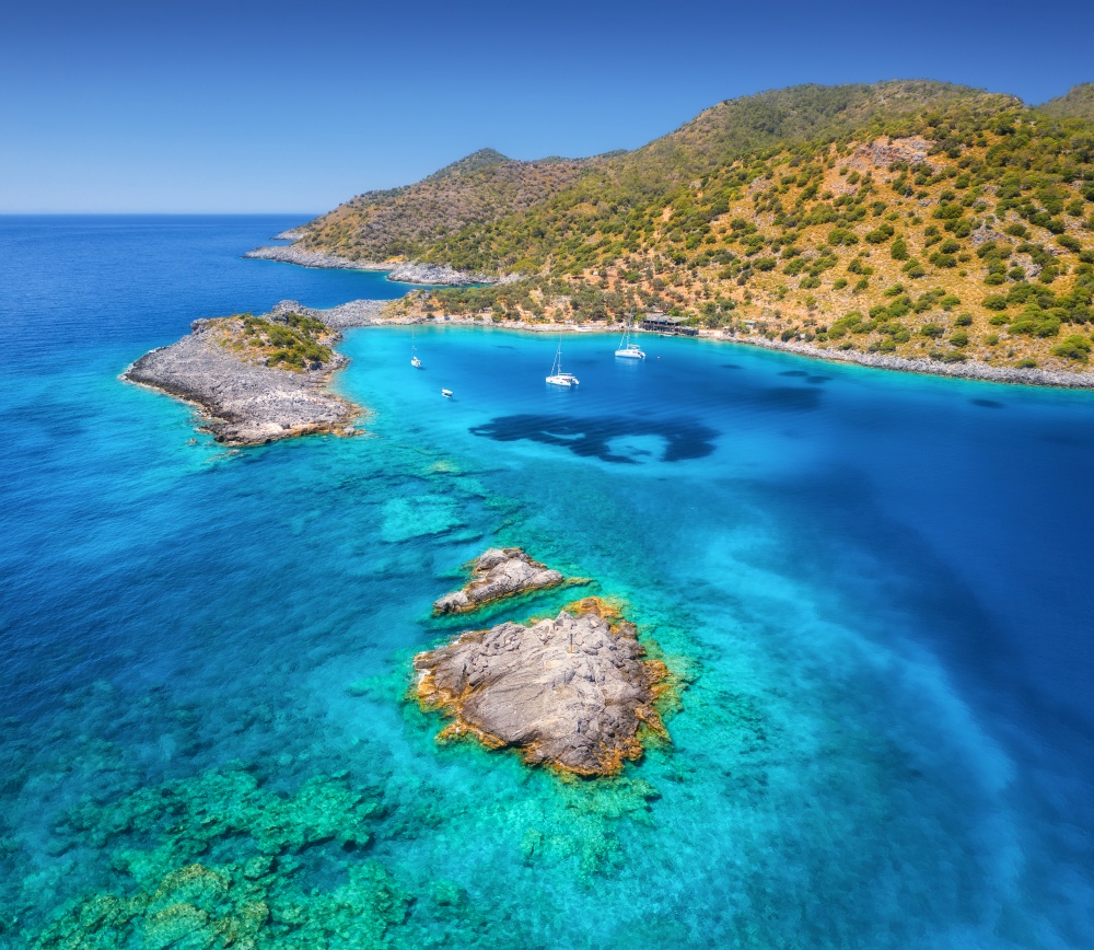 Aerial view of beautiful yachts and boats on the sea at sunny day in summer. Akvaryum koyu in Turkey. Top view of luxury yachts, sailboats, clear blue water, rock, sky, mountain, green trees. Travel