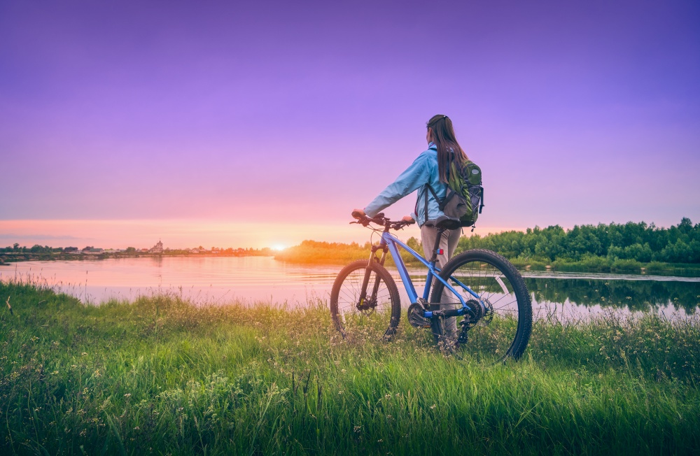 Woman with bicycle near the river at sunset in spring. Landscape with sporty girl with backpack riding a mountain bike, dirt road, green grass, water, purple sky in summer. Sport and travel. Cycle