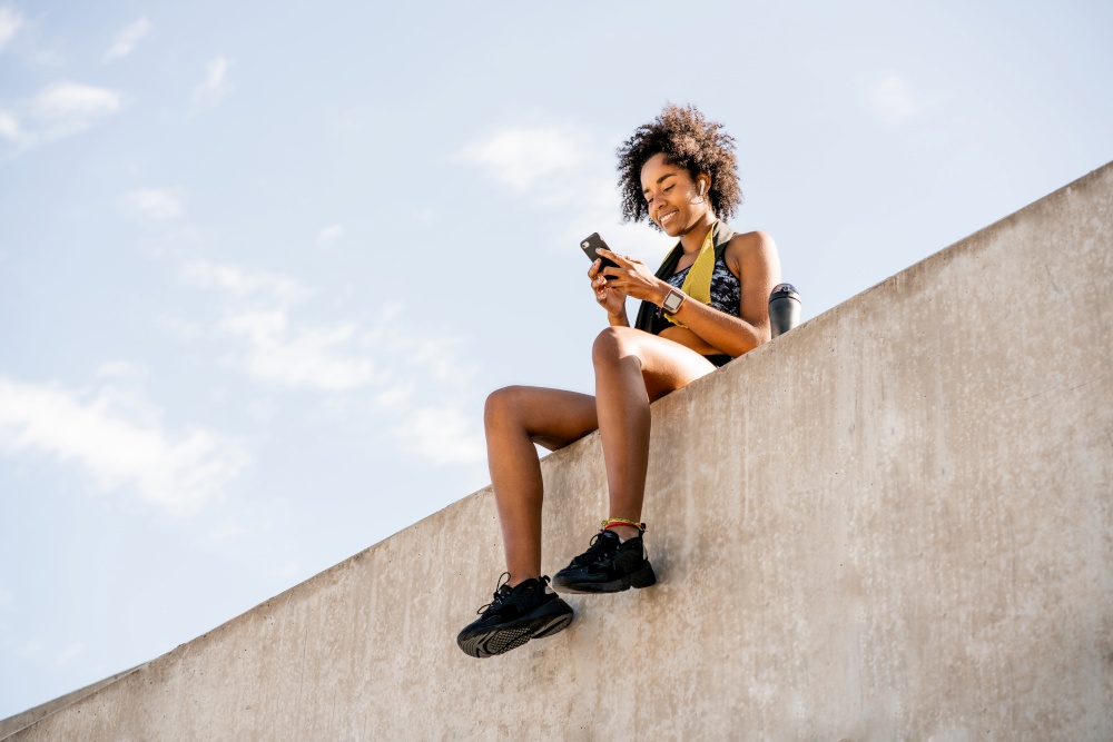 Portrait of afro athlete woman using her mobile phone and relaxing after work out outdoors. Sport and healthy lifestyle.. Athlete woman using her mobile phone outdoors.