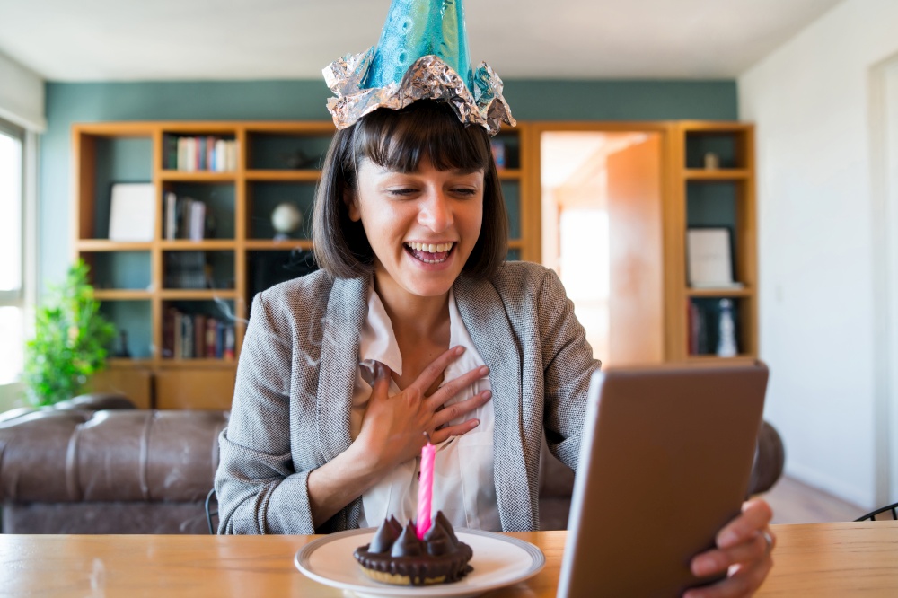 Portrait of young woman celebrating her birthday on a video call with digital tablet and a cake at home. New normal lifestyle concept.. Woman celebrating her birthday on a video call.