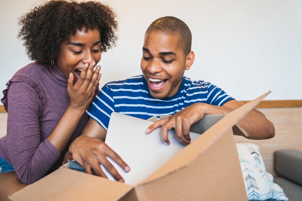 Portrait of happy young latin couple opening a package with a laptop inside, at home. Delivery, shipping and postal service concept.