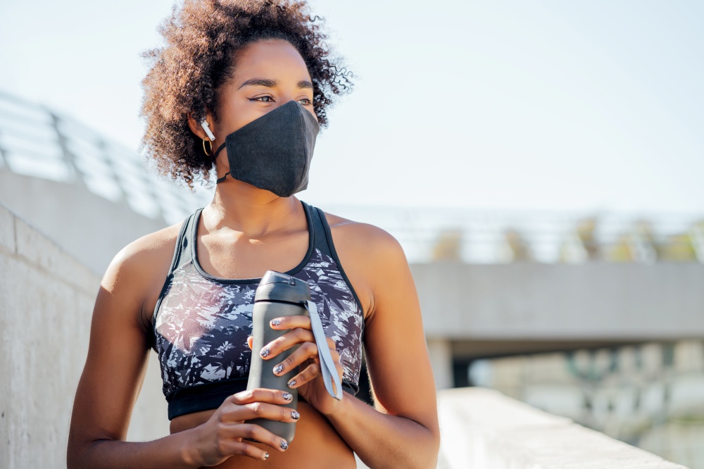 Afro athletic woman wearing face mask and holding a bottle of water after work out outdoors. New normal lifestyle. Sport and healthy lifestyle.