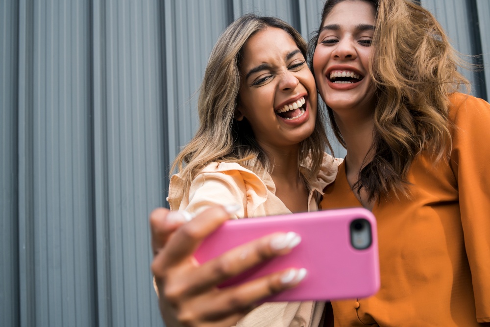 Portrait of two young friends having fun together and taking a selfie with a mobile phone outdoors. Urban concept.