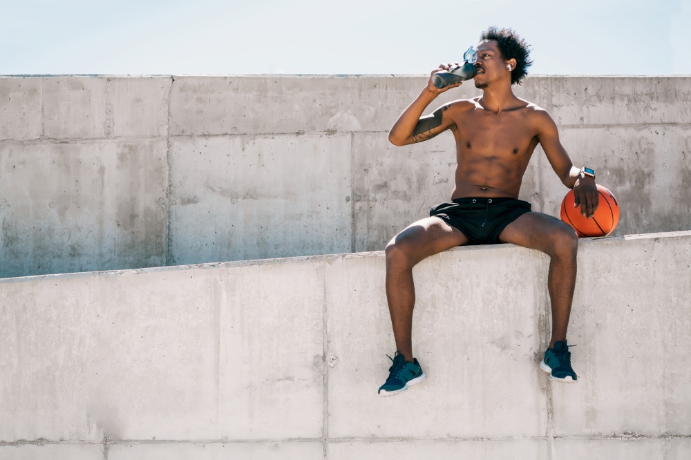 Portrait of afro athlete man drinking water and holding basketball ball outdoors. Sport and healthy lifestyle.