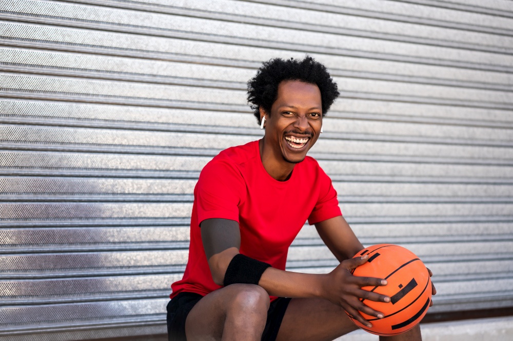 Portrait of afro athlete man holding a basketball ball and relaxing after training while sitting outdoors. Sport and healthy lifestyle.