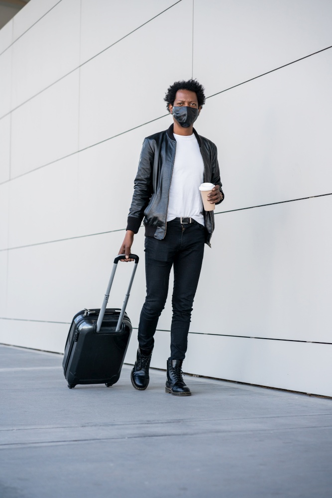 Portrait of afro tourist man carrying suitcase and holding a cup of coffee while walking outdoors on the street. Tourism concept.