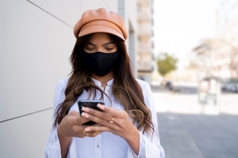 Portrait of young woman wearing protective mask and using her mobile phone while standing outdoors on the street. New normal lifestyle concept. Urban concept.