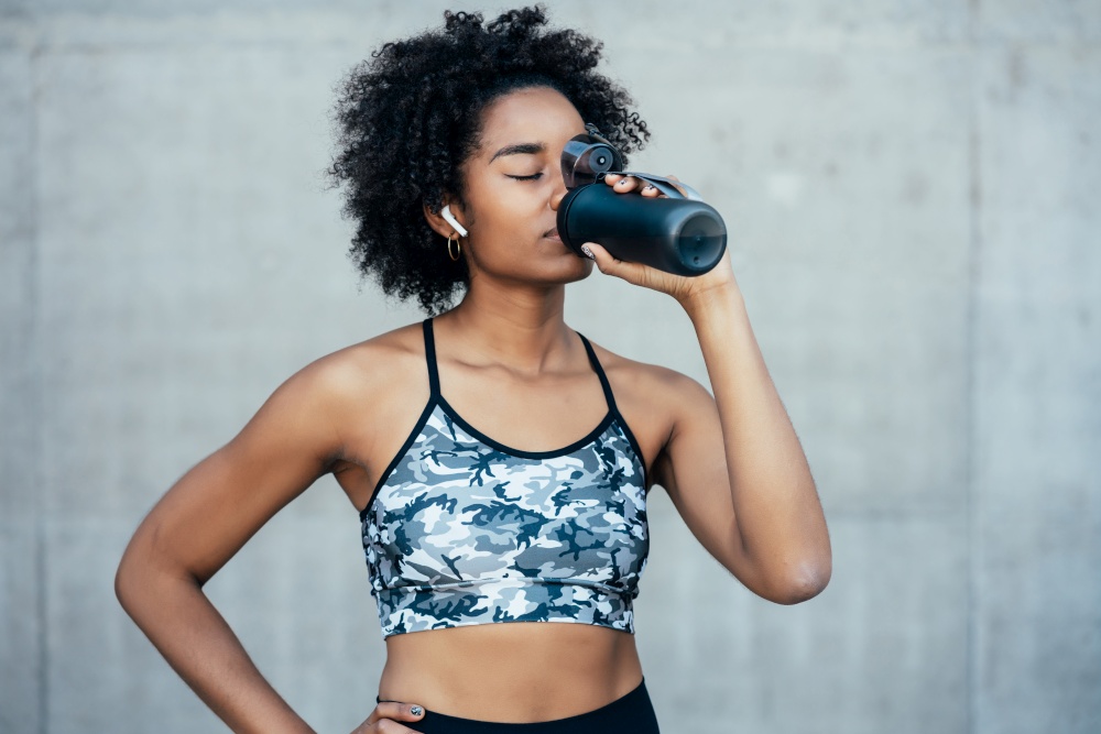 Afro athletic woman drinking water and relaxing after work out outdoors. Sport and healthy lifestyle.