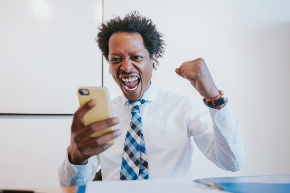 Professional businessman using reading good news on his mobile phone and celebrating victory while working at office. Business and success concept.