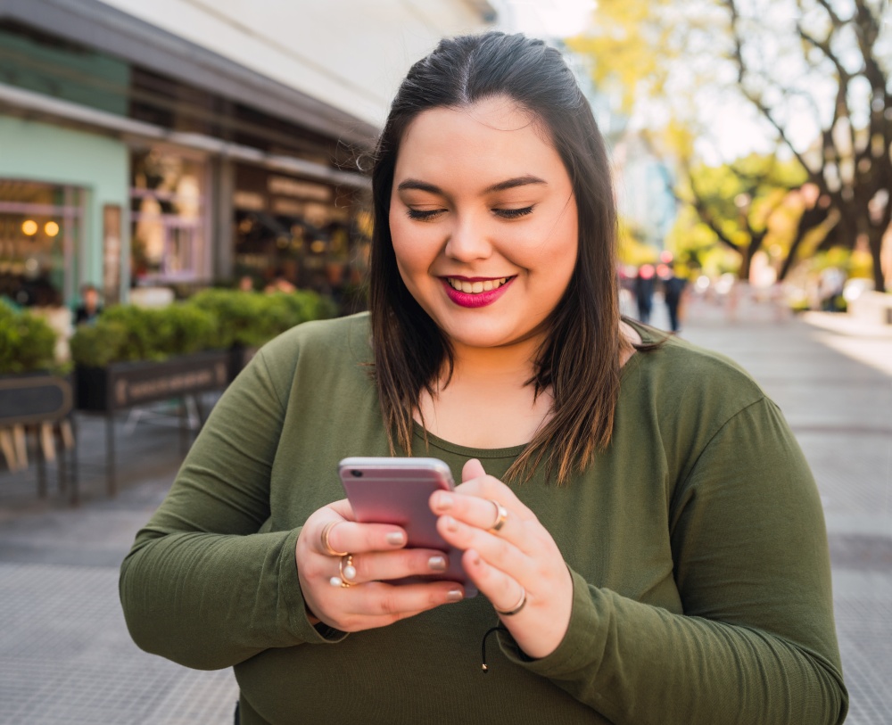 Portrait of young plus size woman typing text message on her mobile phone outdoors on the street. Technology concept.