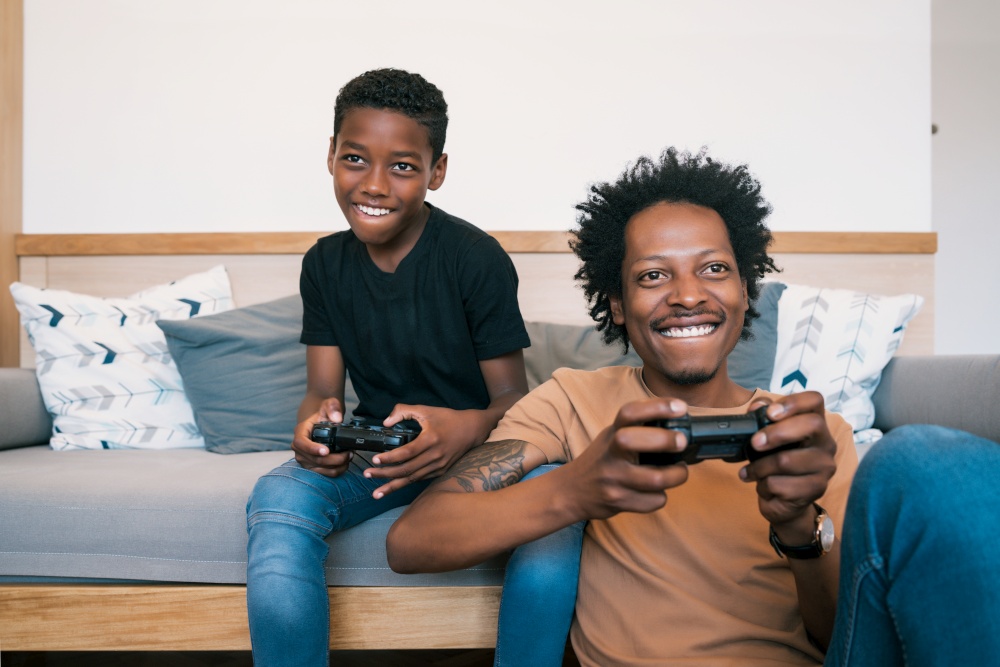 Portrait of happy African American father and son sitting in sofa couch and playing console video games together at home. Family and technology concept.