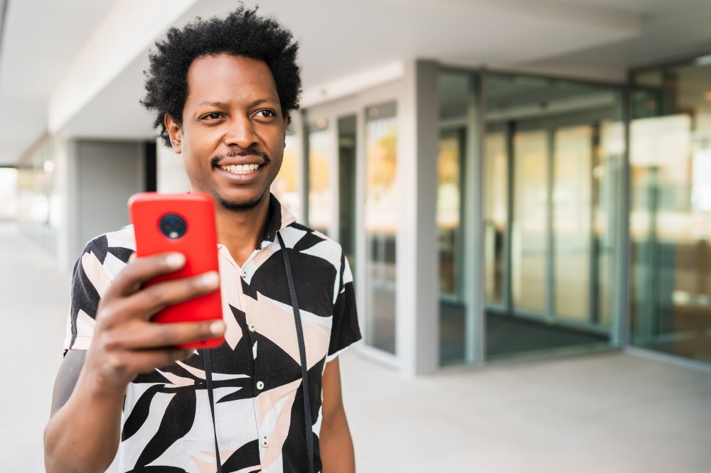 Portrait of afro tourist man using his mobile phone while walking outdoors on the street. Tourism concept.