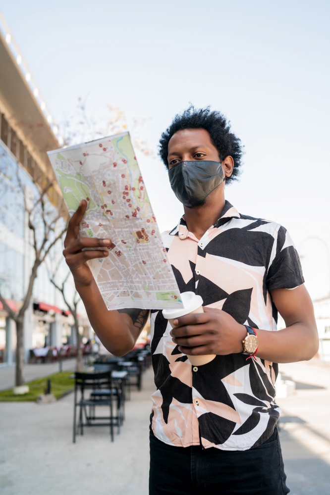 Portrait of afro tourist man wearing protective mask and looking for directions on map while walking outdoors on the street. Tourism concept.