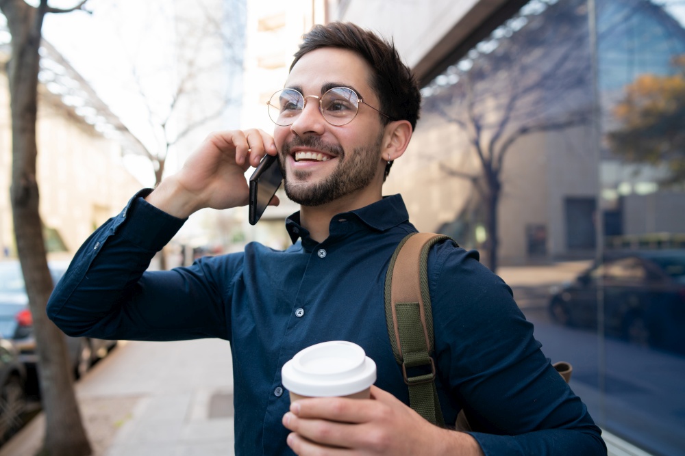 Portrait of young man talking on the phone and holding a cup of coffee while walking outdoors on the street. Urban concept.