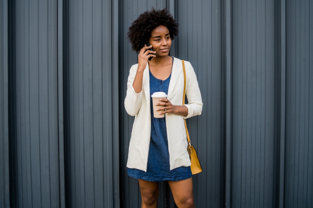 Portrait of afro business woman talking on the phone and holding a cup of coffee while standing outdoors at the street. Business and urban concept.
