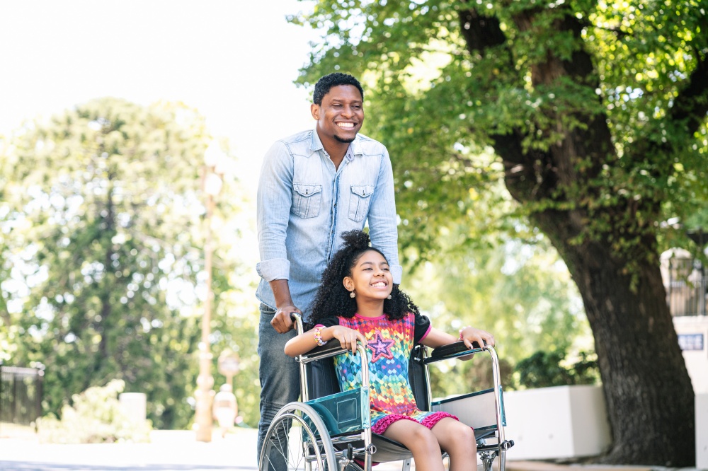 A little girl in a wheelchair enjoying and having fun with her father while on a walk together outdoors.
