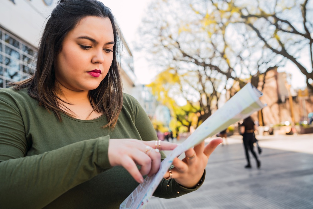 Portrait of young plus size woman holding a map and looking for directions outdoors in the street. Travel concept.