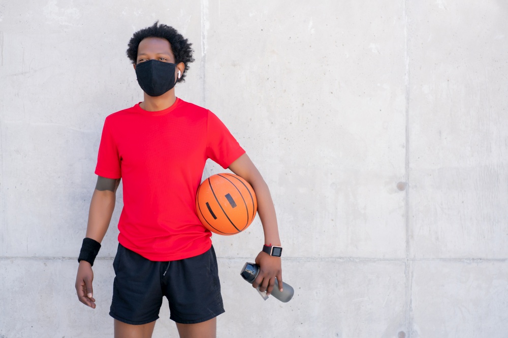 Athletic man wearing face mask and holding a basketball ball while relaxing after training outdoors. New normal lifestyle. Sport and healthy lifestyle concept.