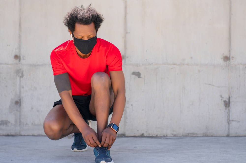 Afro athletic man wearing face mask while tying his shoelaces and getting ready for work out outdoors. New normal lifestyle. Sport and healthy lifestyle concept.