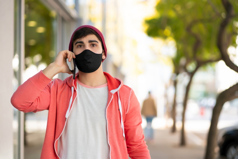 Portrait of young man talking on the phone while walking outdoors on the street. Man wearing face mask. Urban concept.