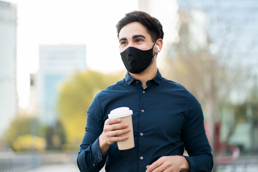 Portrait of young man holding a cup of coffee while standing outdoors on the street. New normal lifestyle concept.