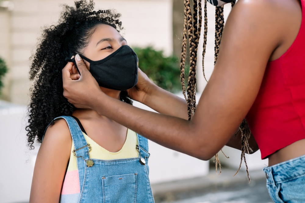 Mother putting a face mask on her daughter while standing outdoors on the street. New normal lifestyle concept.