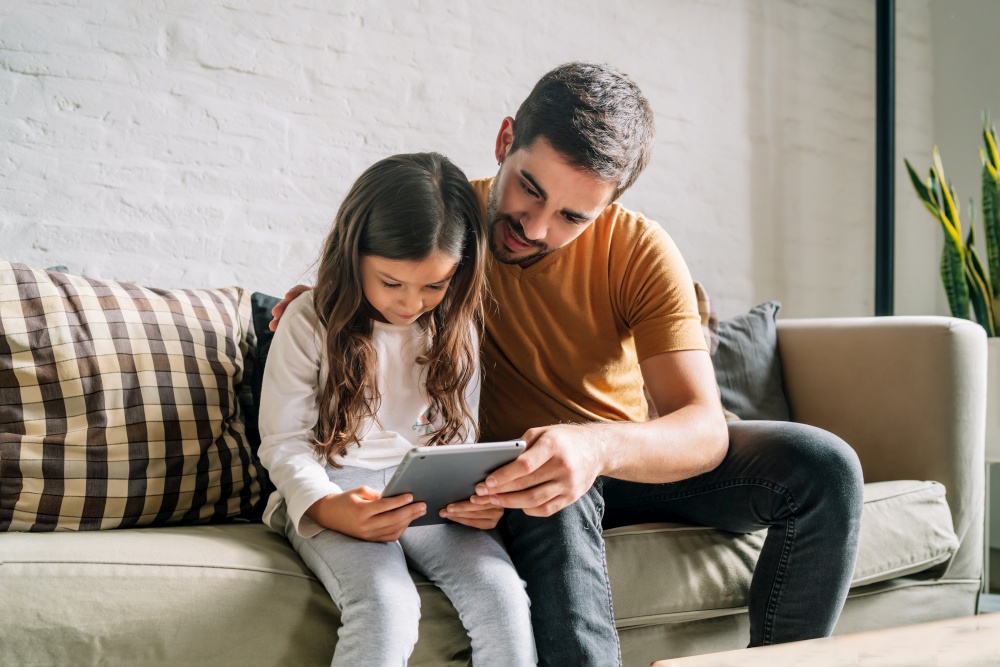 Little girl and her father having fun together while playing with a digital tablet at home. Monoparental family concept.