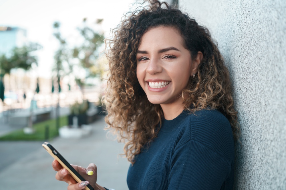 Young latin woman smiling while using her mobile phone outdoors on the street. Urban concept.