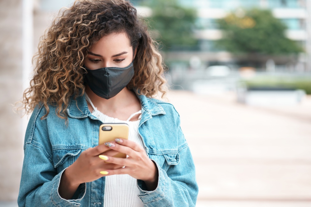 Young latin woman wearing a face mask while using her mobile phone outdoors in the street. New normal lifestyle. Urban concept.