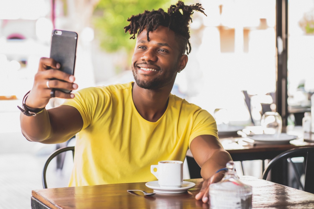 African American man taking a selfie with a mobile phone while sitting at a coffee shop.