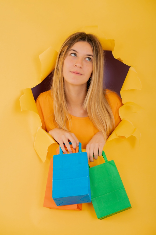 Young woman holding colourful paper bags through a hole in paper wall. Shop concept.