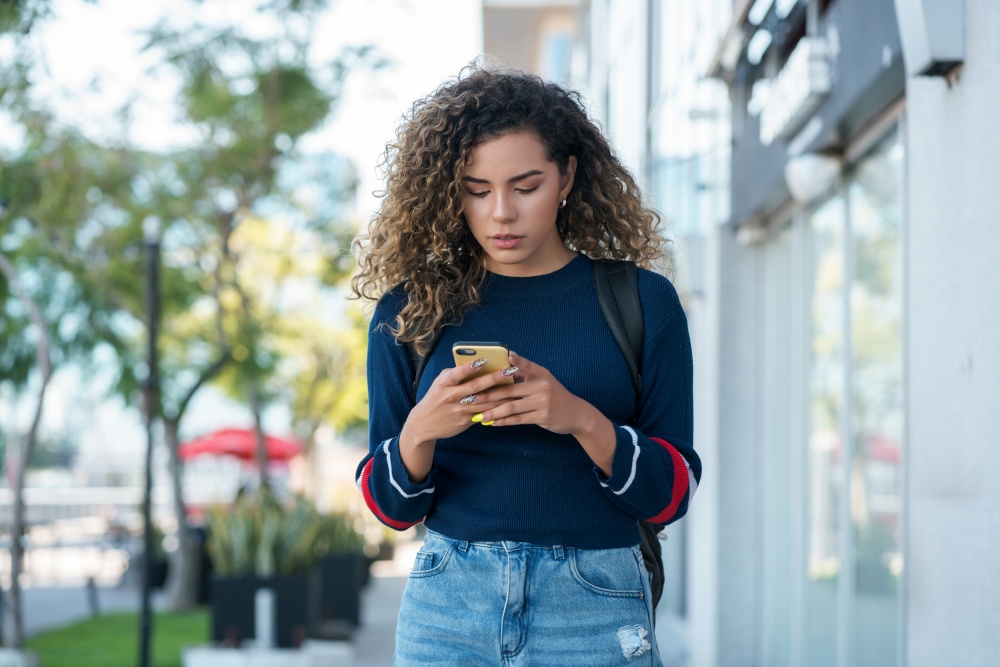 Young latin woman using her mobile phone while walking outdoors on the street. Urban concept.
