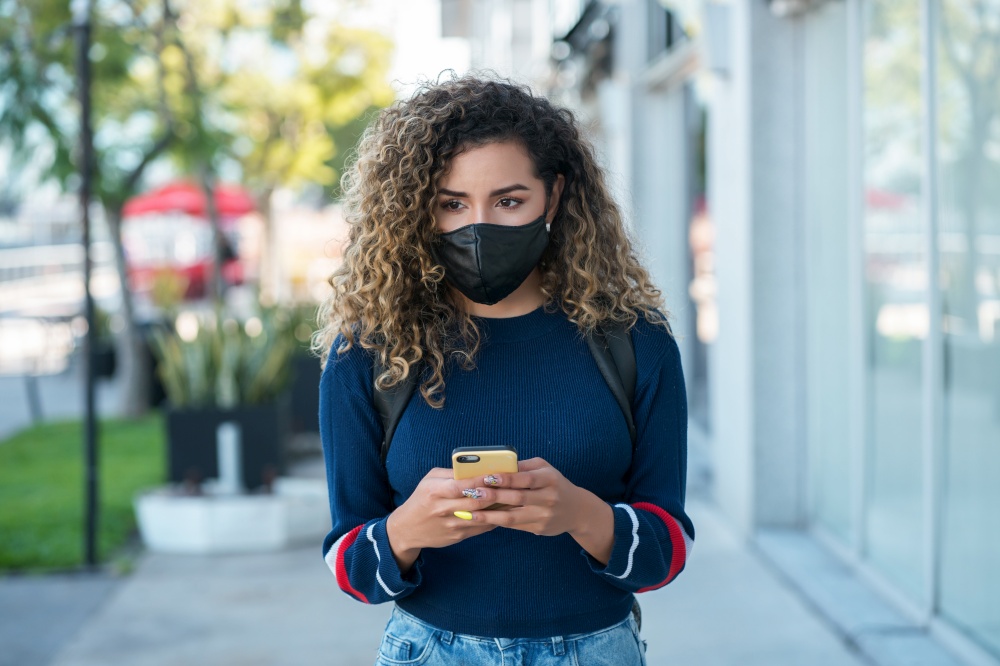 Young latin woman wearing a face mask while using her mobile phone outdoors in the street. New normal lifestyle concept. Urban concept.