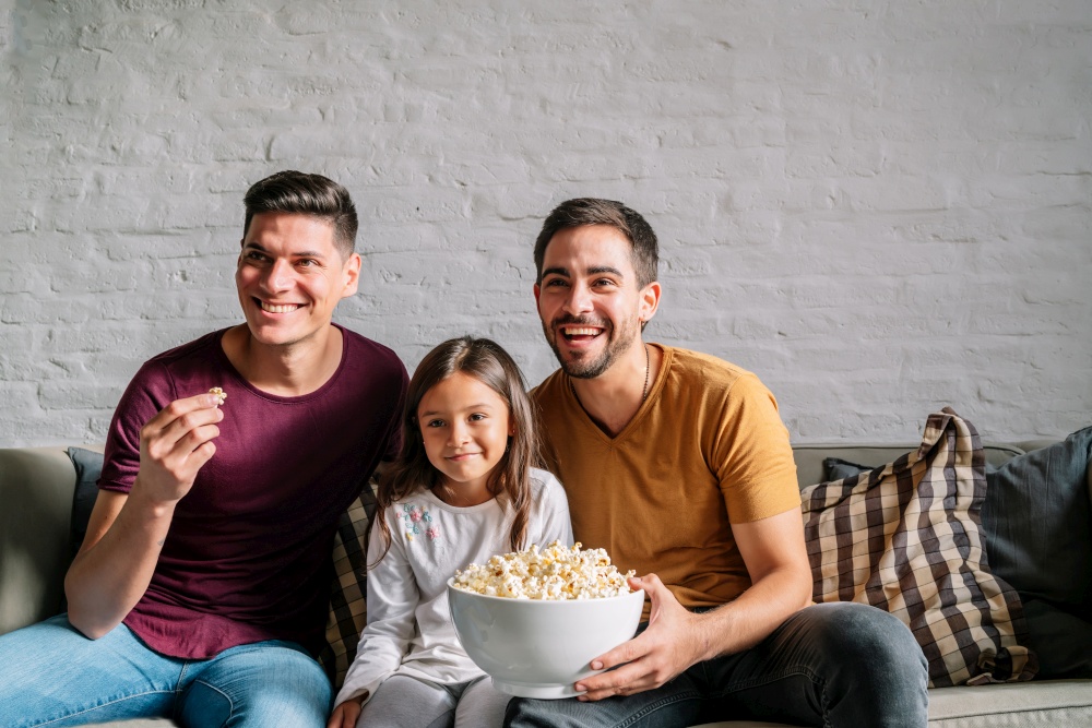 Gay couple enjoying watching movies with their daughter at home. Family concept.