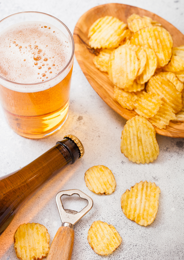 Glass and bottle of craft lager beer with potato crisps snack and opener on stone kitchen background. Beer and snack.